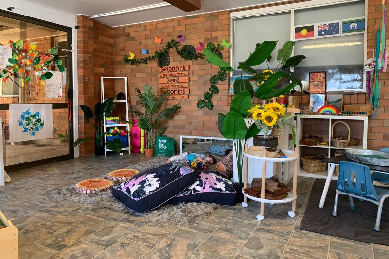 A cozy, colorful classroom corner with cushions, soft toys, and educational decorations. A bulletin board displays messages and art. Large plants and sunflowers add a natural touch, while shelves hold books and toys for children.