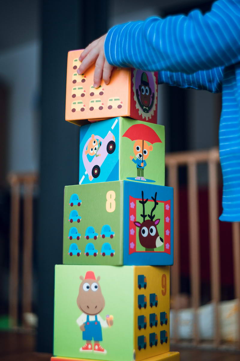 A child wearing a blue striped shirt is stacking colorful toy blocks at Excellence In Care family day care. The blocks, adorned with cartoon animals and numbers, form an impressive vertical tower.